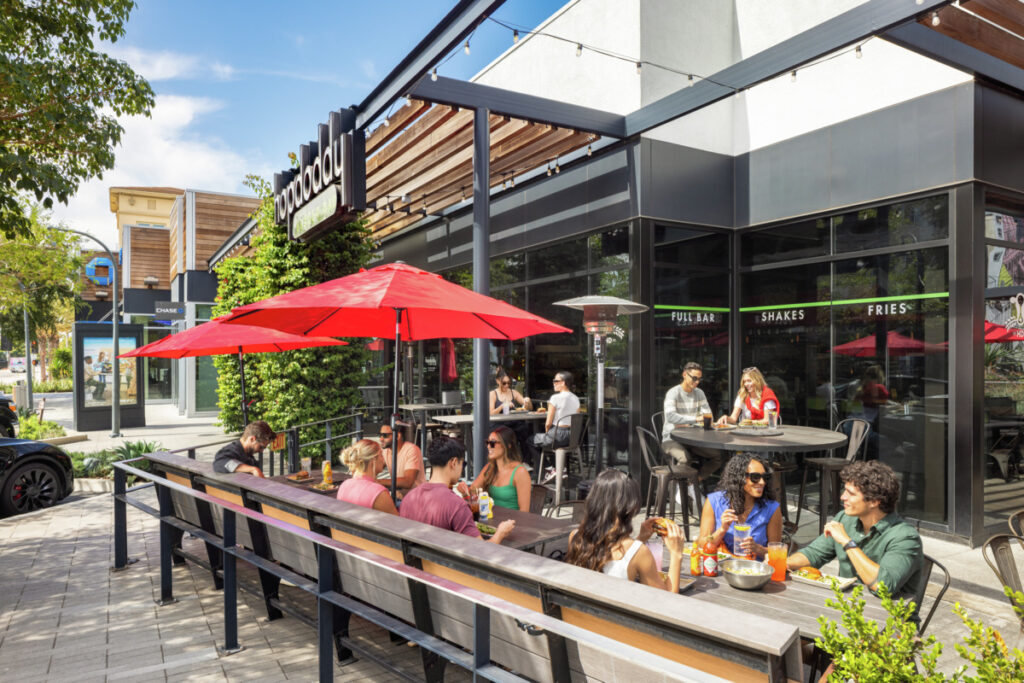 Photo rendering of the outdoor shops at Runway featuring multiple people dining outside on a wooden bench under a red umbrella. 