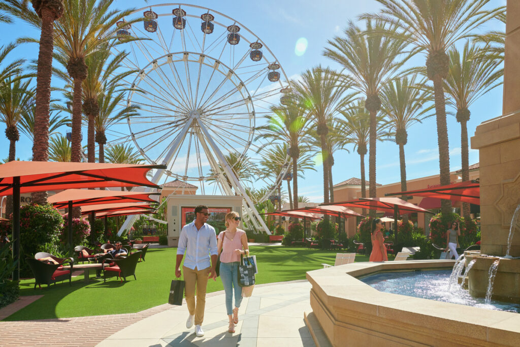 Front view photo rendering of a man and woman holding hands strolling through Irvine Spectrum park with Ferris wheel behind them and water fountain beside them. 