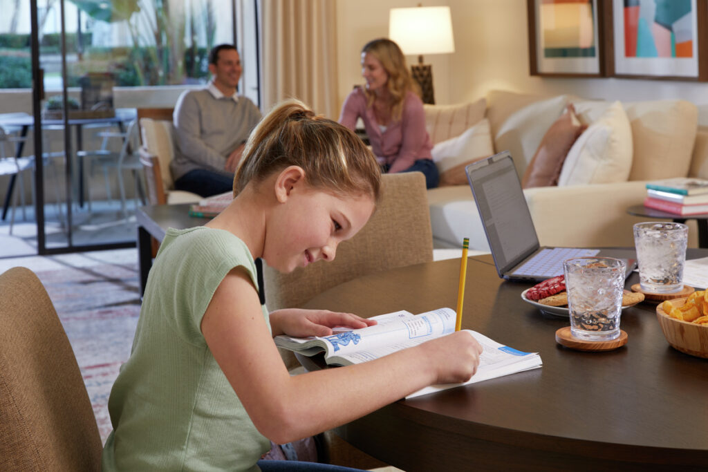Photo of family sitting inside the living room of apartment with parents on the couch and daughter working on schoolwork at a table in the forefront.